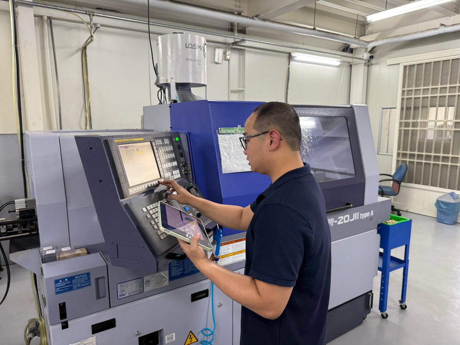 An operator standing in a factory, using a tablet while operating the control panel of a blue Star SR-20JII CNC lathe machine.